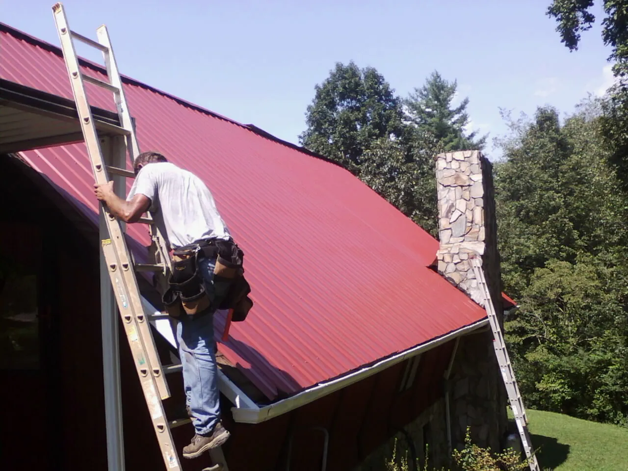 Roofer on a ladder beside a red metal roof and stone chimney — Soffit & Fascia Repair by Roofing Brothers in Weatherly